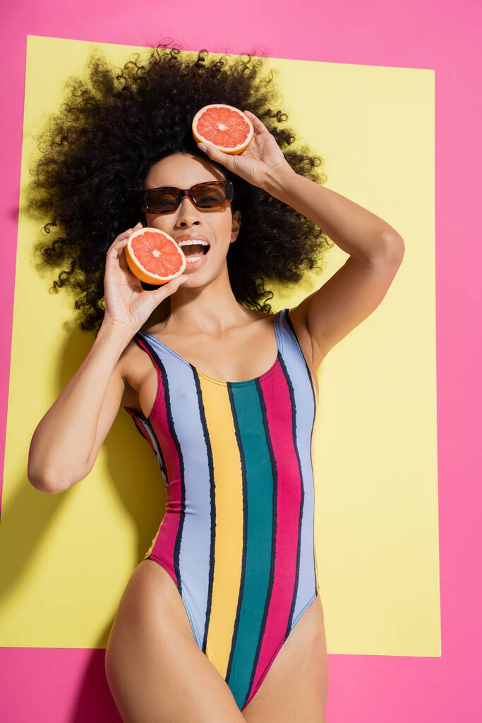 vista superior de la mujer americana africana rizada en gafas de sol y traje de baño colorido mordiendo pomelo sobre fondo amarillo y rosa  - Foto, Imagen