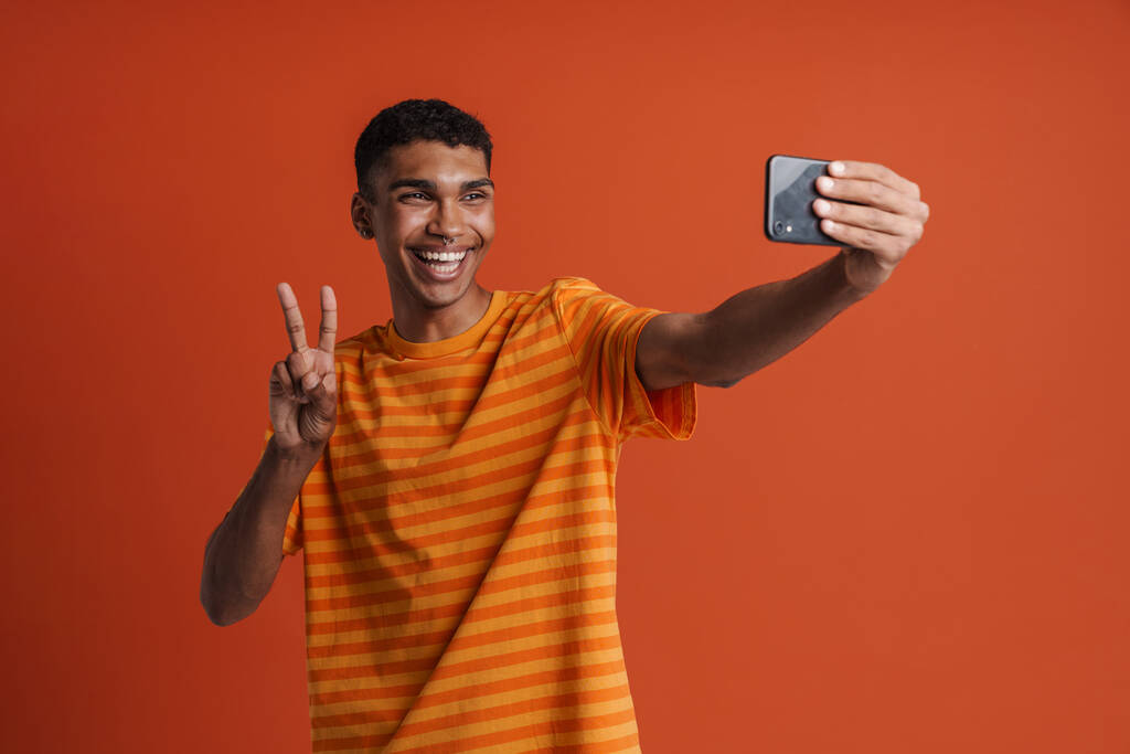 Nuori komea hymyilevä onnellinen afrikkalainen mies lävistyksiä ottaen selfie puhelimessa ja osoittaa voiton ele, kun seisoo yli eristetty oranssi tausta - Valokuva, kuva
