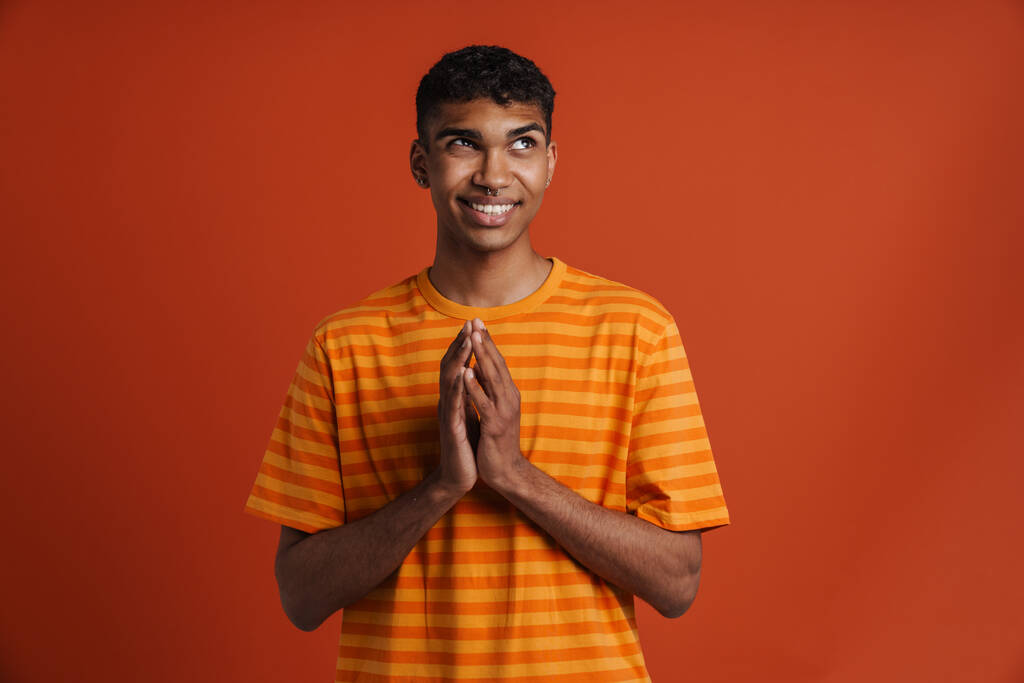 Mladý černošky muž s pronikavým úsměvem při modlitbě gesto izolované přes červené pozadí - Fotografie, Obrázek