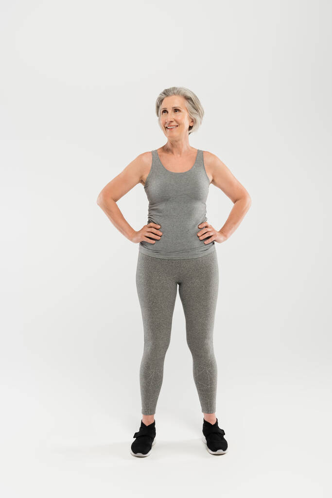 longitud completa de la mujer feliz y mayor en ropa deportiva de pie con las manos en las caderas en gris - Foto, imagen