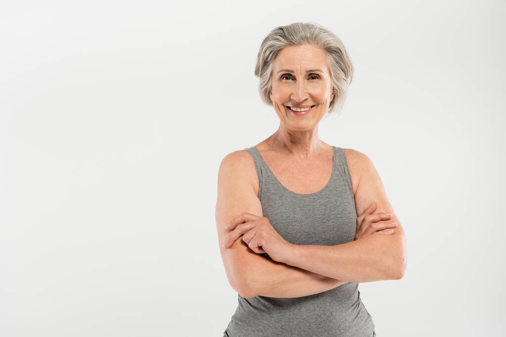 щаслива і старша жінка в спортивному одязі, що стоїть зі складеними руками ізольовані на сірому
 - Фото, зображення