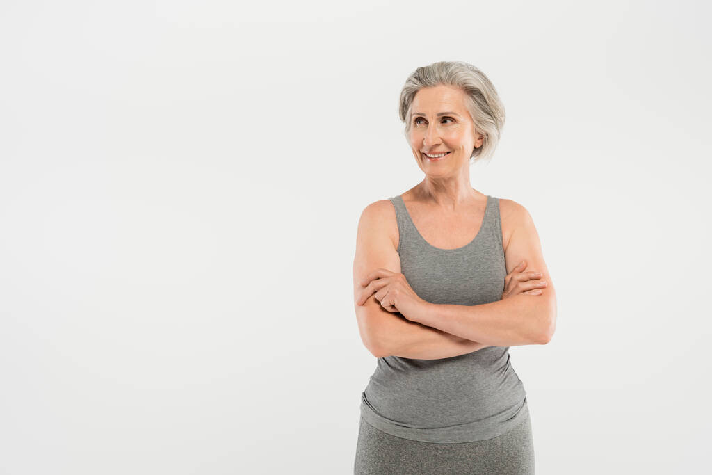 donna allegra e anziana in abbigliamento sportivo in piedi con braccia incrociate isolate su grigio - Foto, immagini