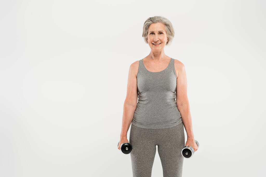 mujer mayor feliz con pelo gris haciendo ejercicio con pesas aisladas en gris  - Foto, imagen