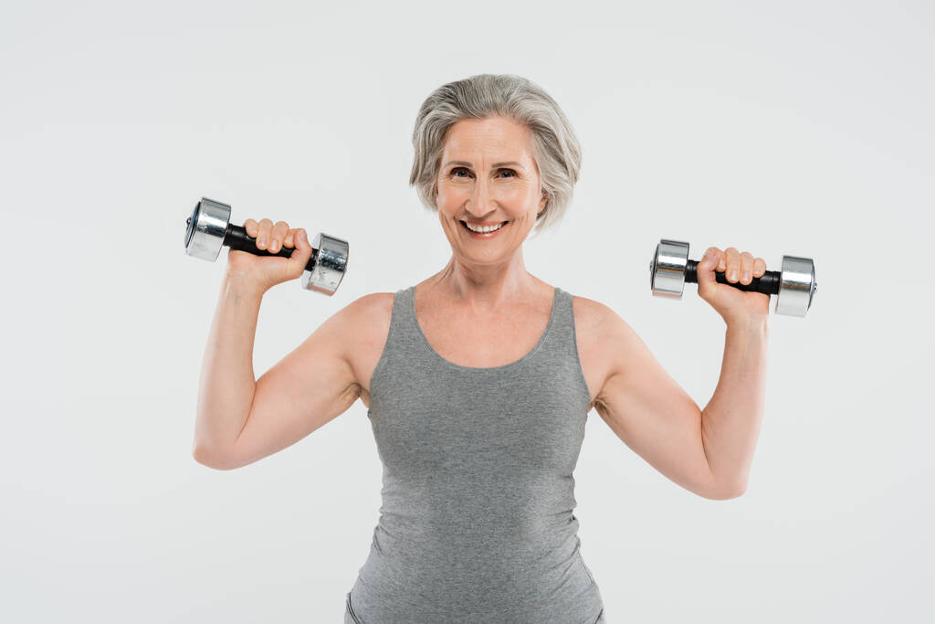ενθουσιασμένοι ηλικιωμένη γυναίκα με γκρίζα μαλλιά άσκηση με αλτήρες απομονώνονται σε γκρι  - Φωτογραφία, εικόνα