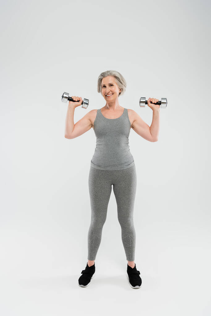 volledige lengte van gelukkig senior vrouw in strakke sportkleding oefening met halters op grijs  - Foto, afbeelding