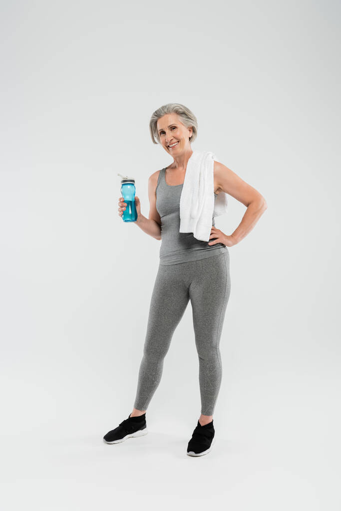 longitud completa de la mujer mayor alegre con toalla blanca en el hombro sosteniendo botella deportiva con agua en gris  - Foto, imagen