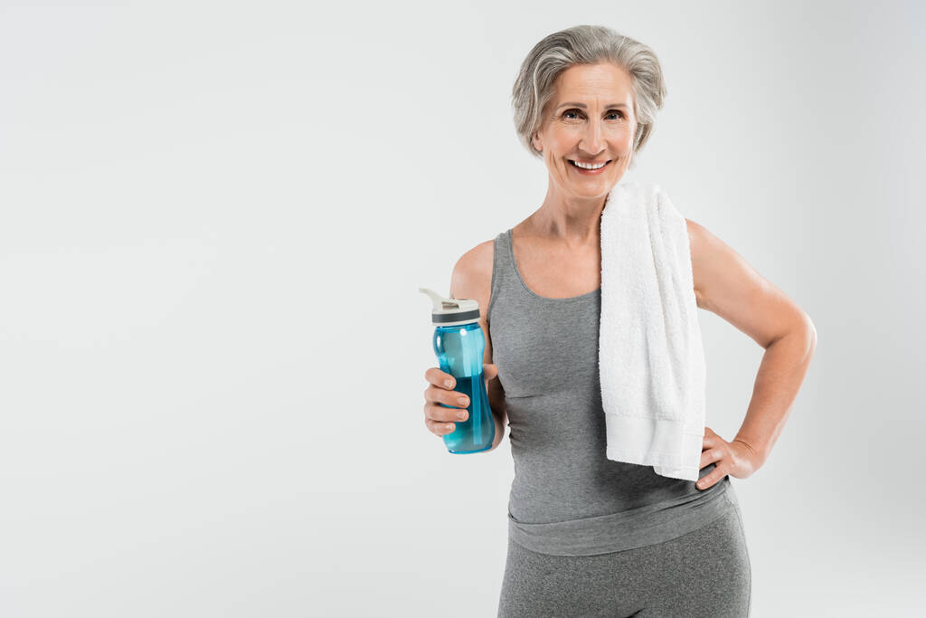 mujer mayor llena de alegría con toalla blanca en el hombro sosteniendo botella deportiva con agua aislada en gris  - Foto, imagen
