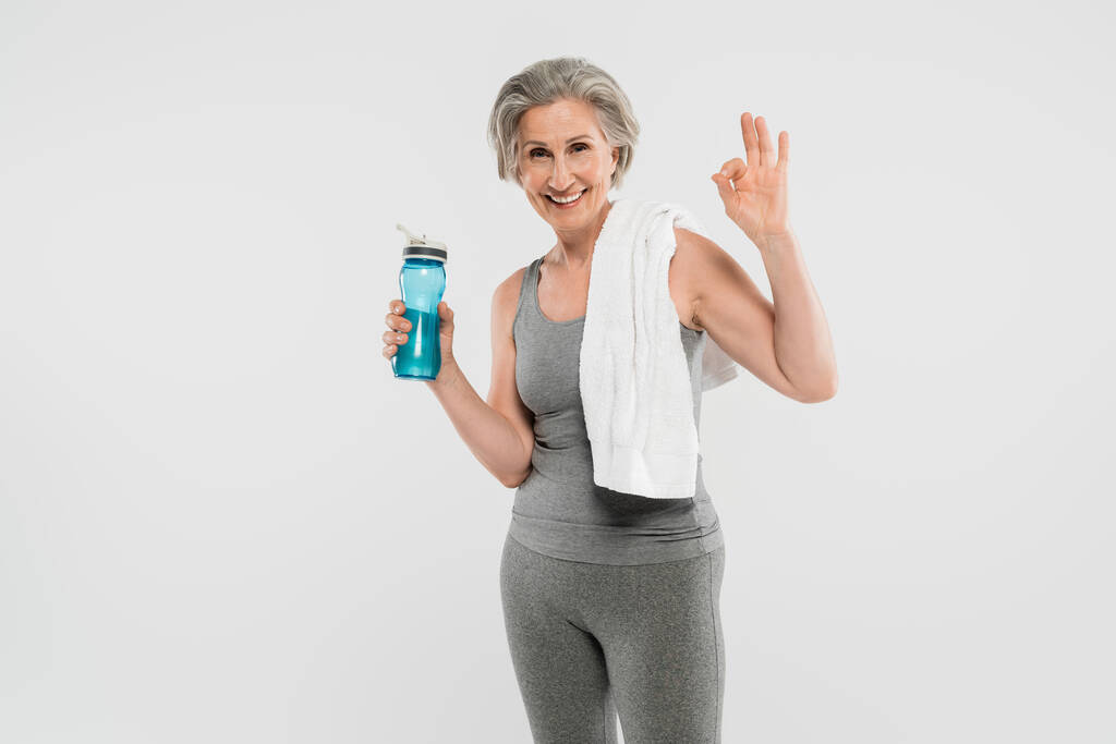 ευτυχισμένη ηλικιωμένη γυναίκα με λευκή πετσέτα στον ώμο κρατώντας αθλητικό μπουκάλι με νερό και δείχνει ok απομονώνονται σε γκρι  - Φωτογραφία, εικόνα