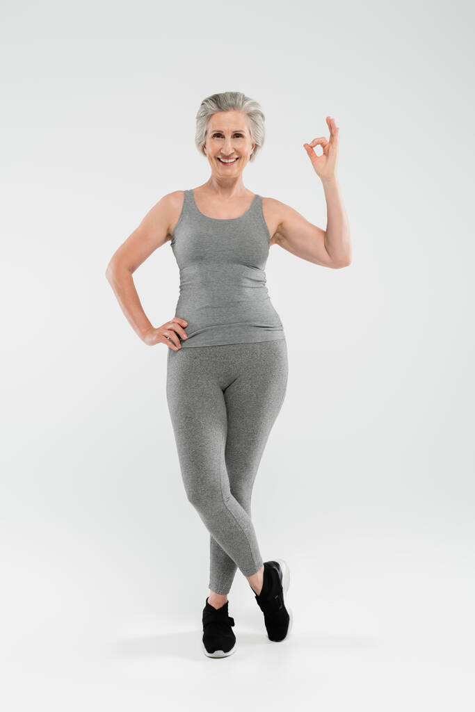 longitud completa de mujer alegre y jubilada en ropa deportiva posando con la mano en la cadera y mostrando bien en gris  - Foto, imagen