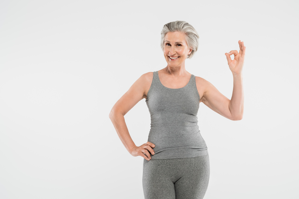 mulher feliz e aposentado em sportswear posando com a mão no quadril e mostrando bem isolado em cinza - Foto, Imagem