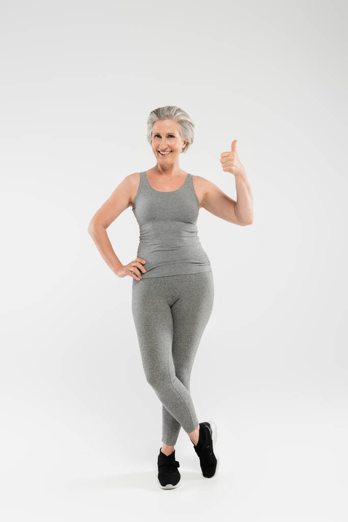 full length of cheerful and retired woman in sportswear posing with hand on hip and showing like on grey  - Photo, Image