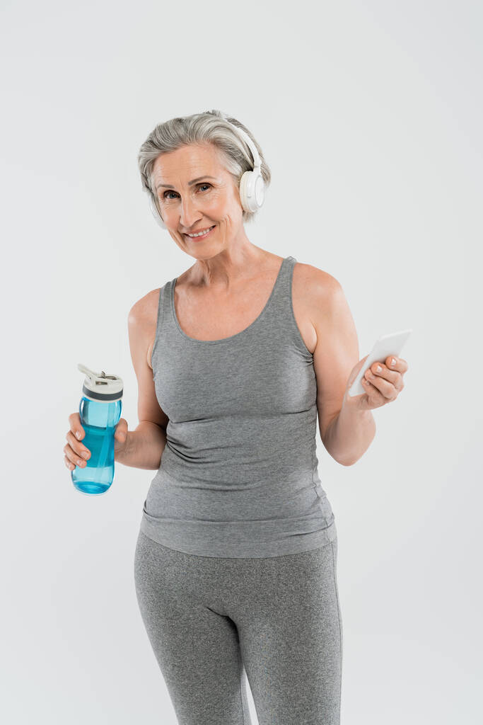 fröhliche Seniorin hört Musik in drahtlosen Kopfhörern, während sie Sportflasche und Smartphone isoliert auf grau hält  - Foto, Bild