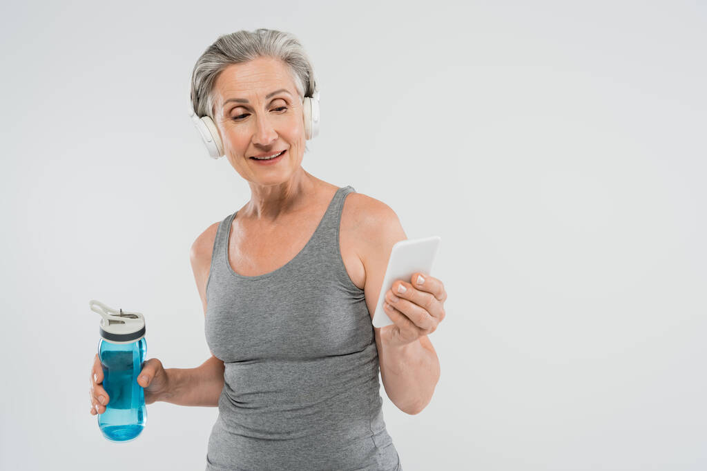 щаслива старша жінка слухає музику в бездротових навушниках, тримаючи спортивну пляшку і смартфон ізольовано на сірому
  - Фото, зображення