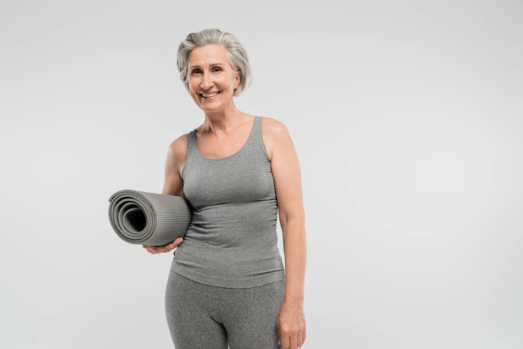 bezstarostná starší žena ve sportovním oblečení drží fitness rohožku a usmívá se izolovaně na šedé  - Fotografie, Obrázek