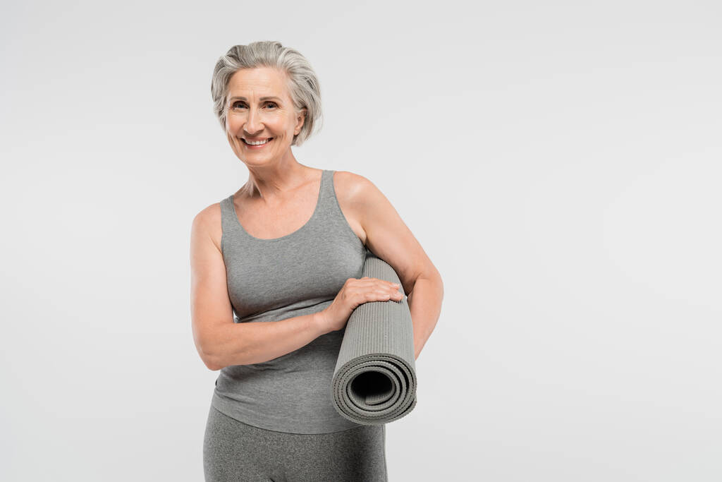 Χαρούμενη ηλικιωμένη γυναίκα σε αθλητικά κρατώντας στρώμα γυμναστικής και χαμογελώντας απομονωμένη σε γκρι  - Φωτογραφία, εικόνα