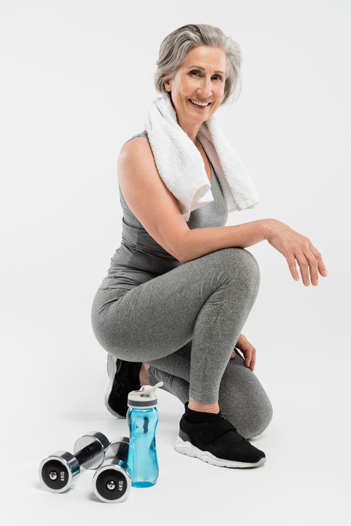 full length of happy senior woman with towel on shoulder smiling while sitting on grey  - Photo, Image
