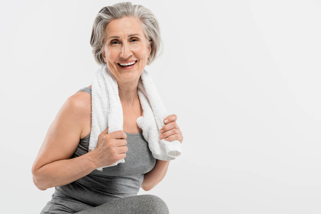 glückliche Seniorin mit weißem Handtuch und Blick auf Kamera isoliert auf grau  - Foto, Bild