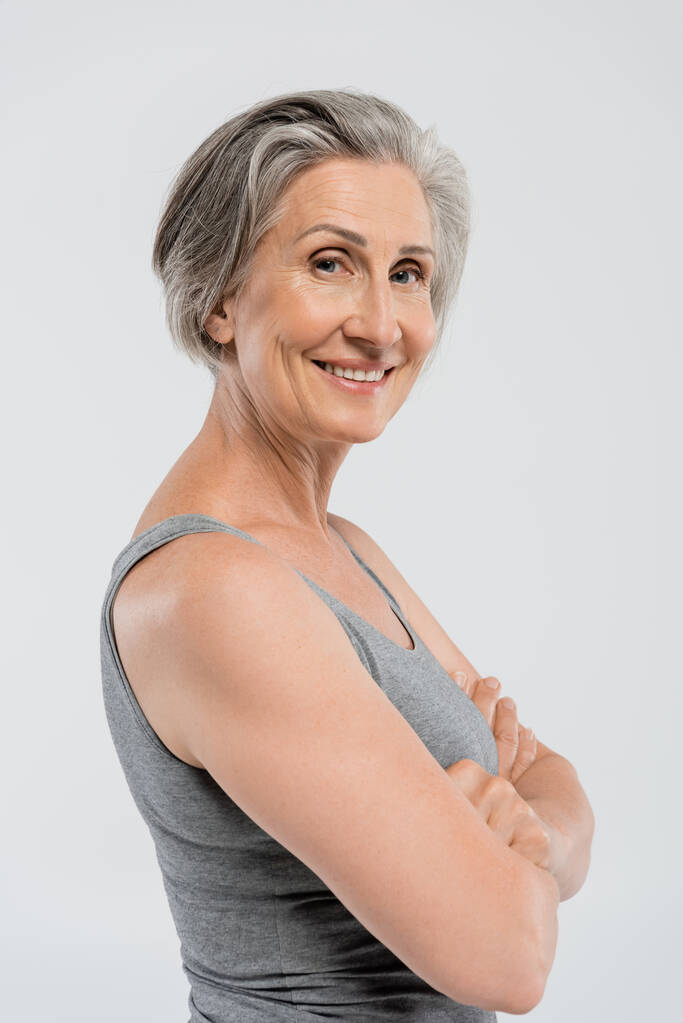 mulher sênior feliz com cabelos grisalhos de pé com braços cruzados isolados em cinza  - Foto, Imagem