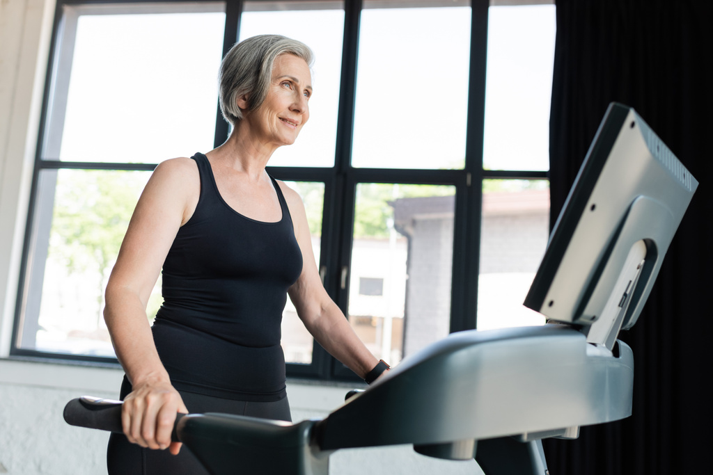 Fröhliche Seniorin mit grauen Haaren läuft im Fitnessstudio auf Laufband  - Foto, Bild