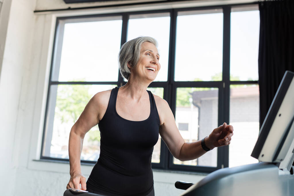насолоджена старша жінка з сірим волоссям біжить на біговій доріжці в спортзалі
  - Фото, зображення