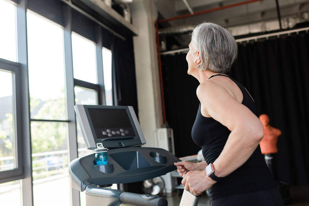 retired woman with grey hair running on treadmill next to sports bottle with water  - Photo, Image