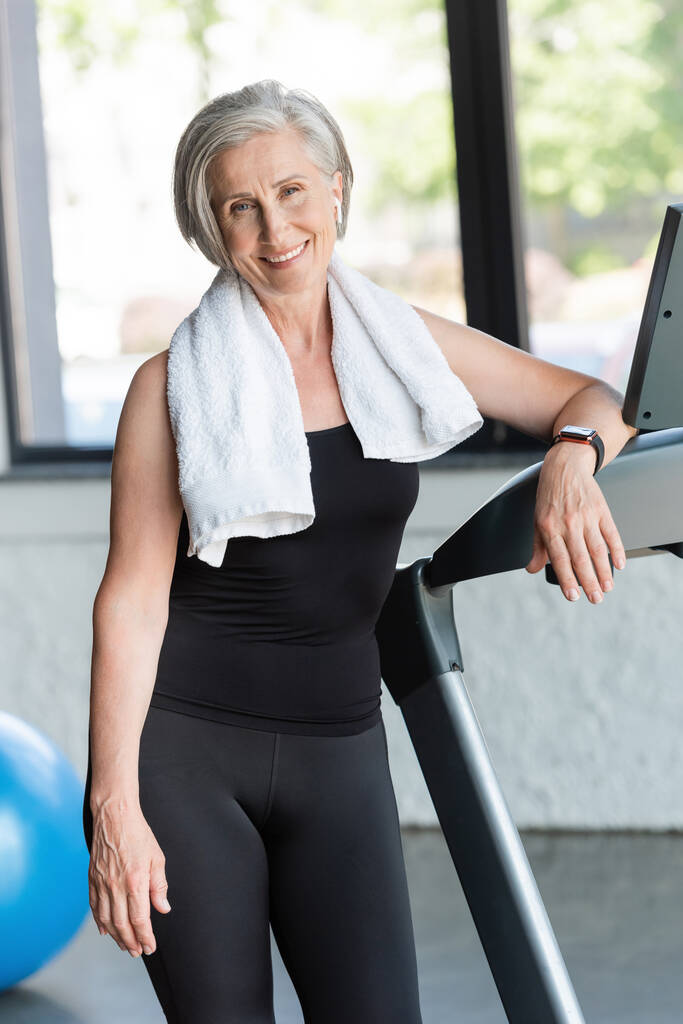 joyful senior woman with white towel on shoulders standing next to treadmill  - Photo, Image