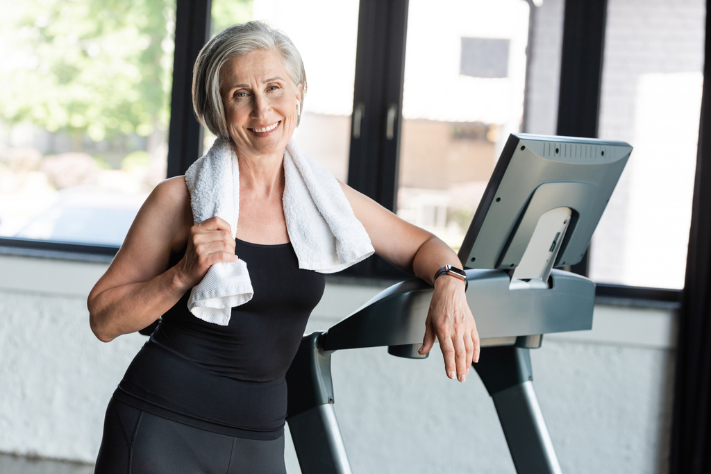 joyful retired woman with white towel on shoulders standing next to treadmill  - Photo, Image