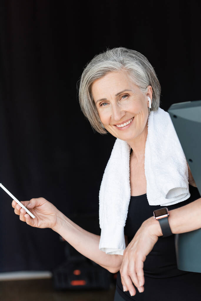 veselá a starší žena v bezdrátovém sluchátku pomocí smartphone v tělocvičně  - Fotografie, Obrázek