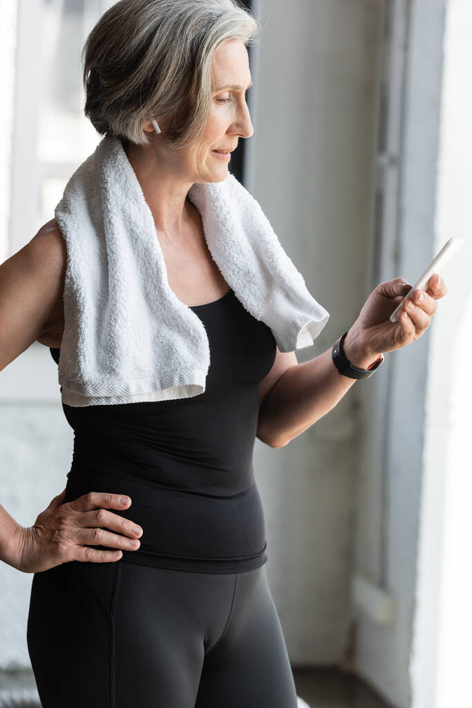vejde starší žena s ručníkem na ramena pomocí smartphone při poslechu hudby v bezdrátovém sluchátku  - Fotografie, Obrázek