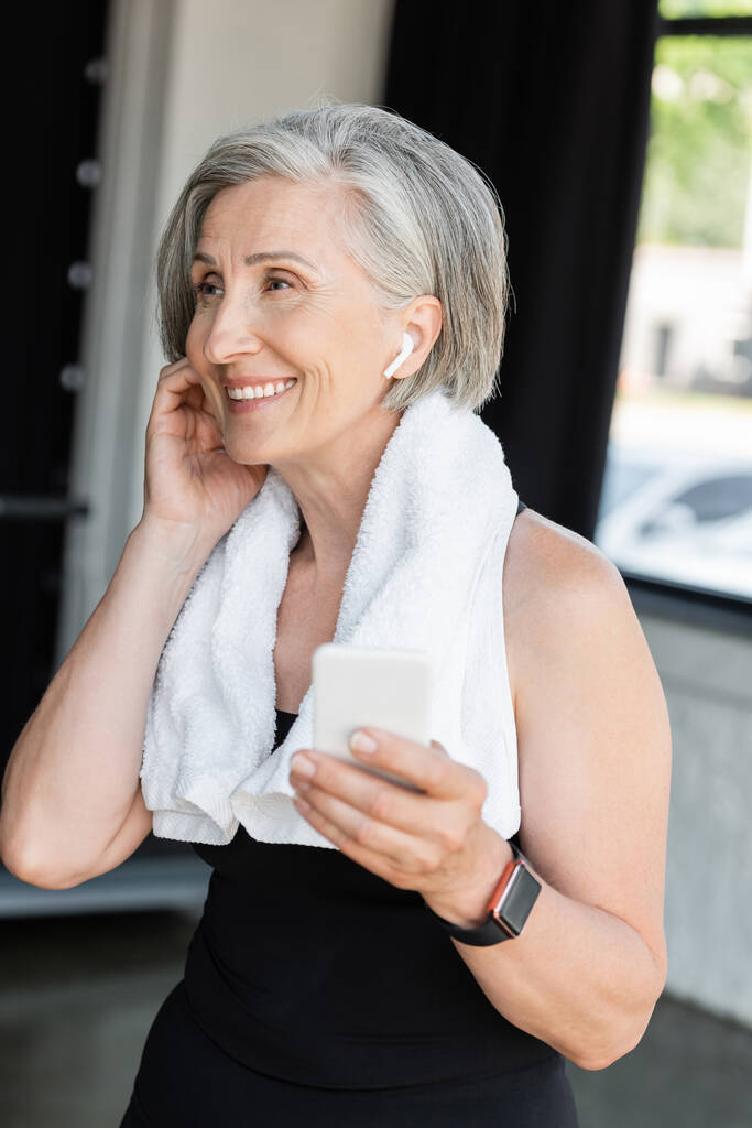 szczęśliwa starsza kobieta z ręcznikiem na ramionach za pomocą smartfona podczas noszenia słuchawki bezprzewodowej  - Zdjęcie, obraz