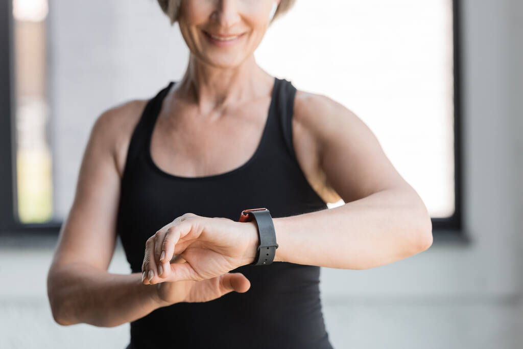 cropped view of cheerful senior woman in black tank top checking activity on fitness tracker in gym  - Photo, Image