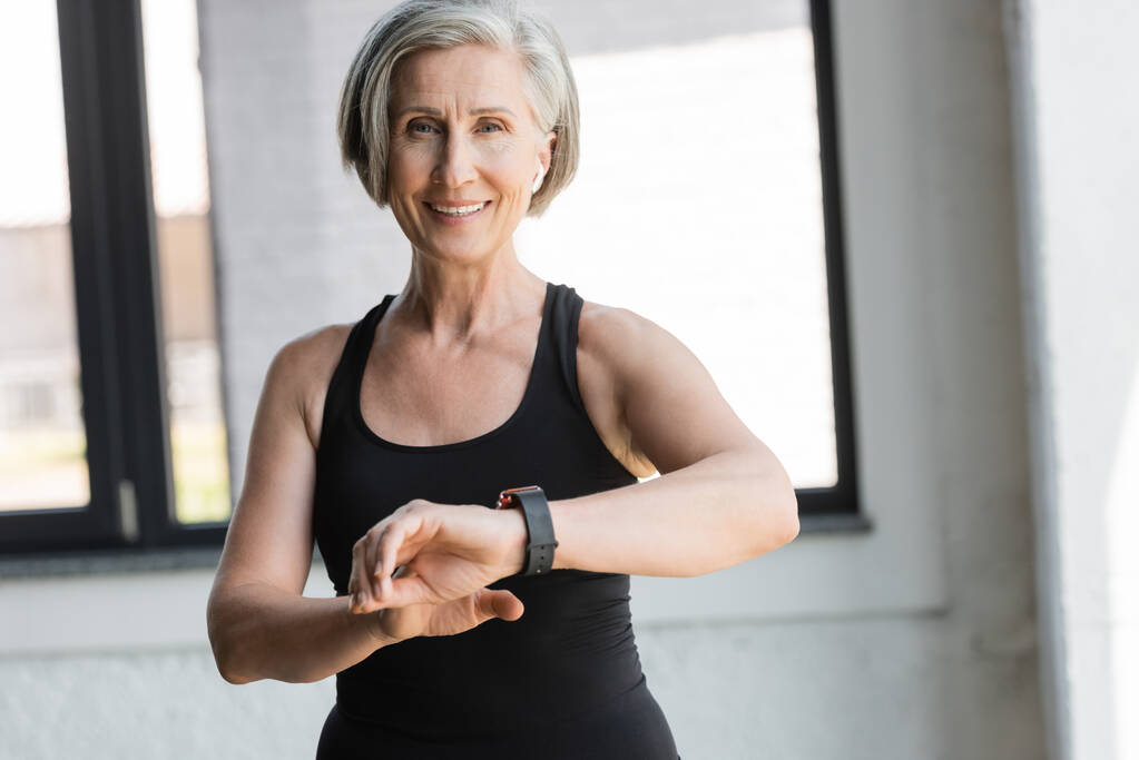 vejde starší žena v černé tílko s úsměvem při kontrole aktivity na fitness tracker v tělocvičně  - Fotografie, Obrázek
