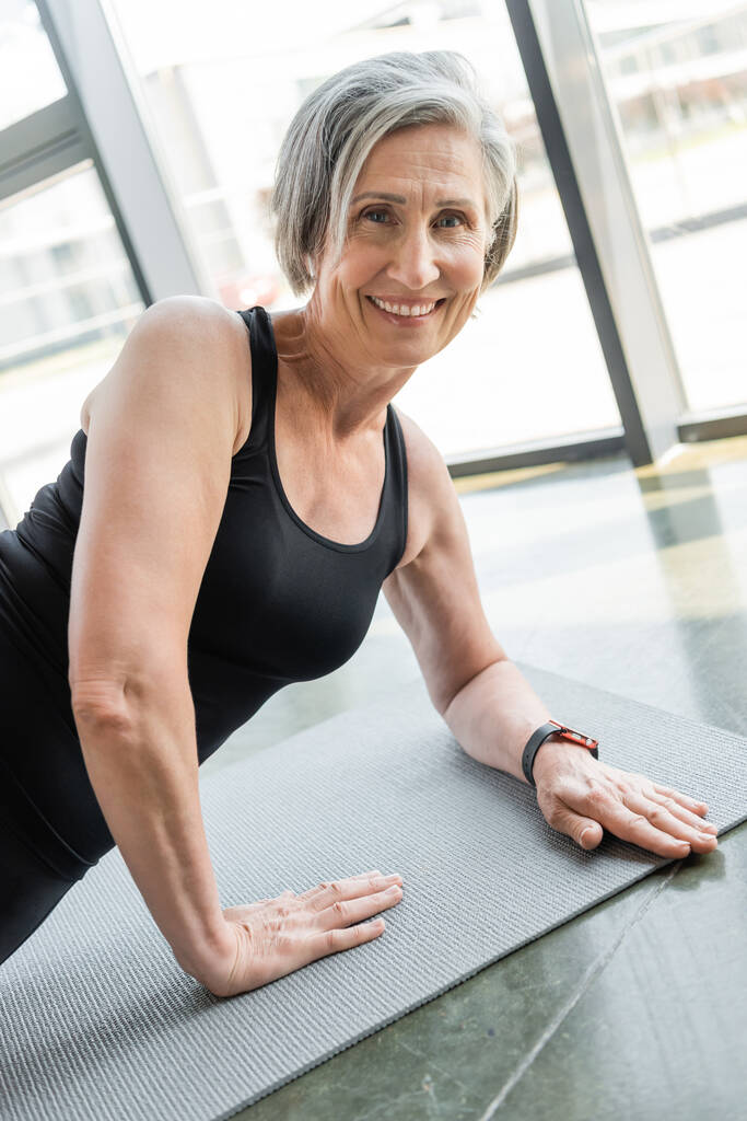 senior sportswoman smiling while exercising on fitness mat in gym  - Photo, Image