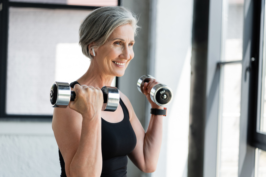 happy and senior woman in wireless earphone exercising with dumbbells in gym  - Photo, Image