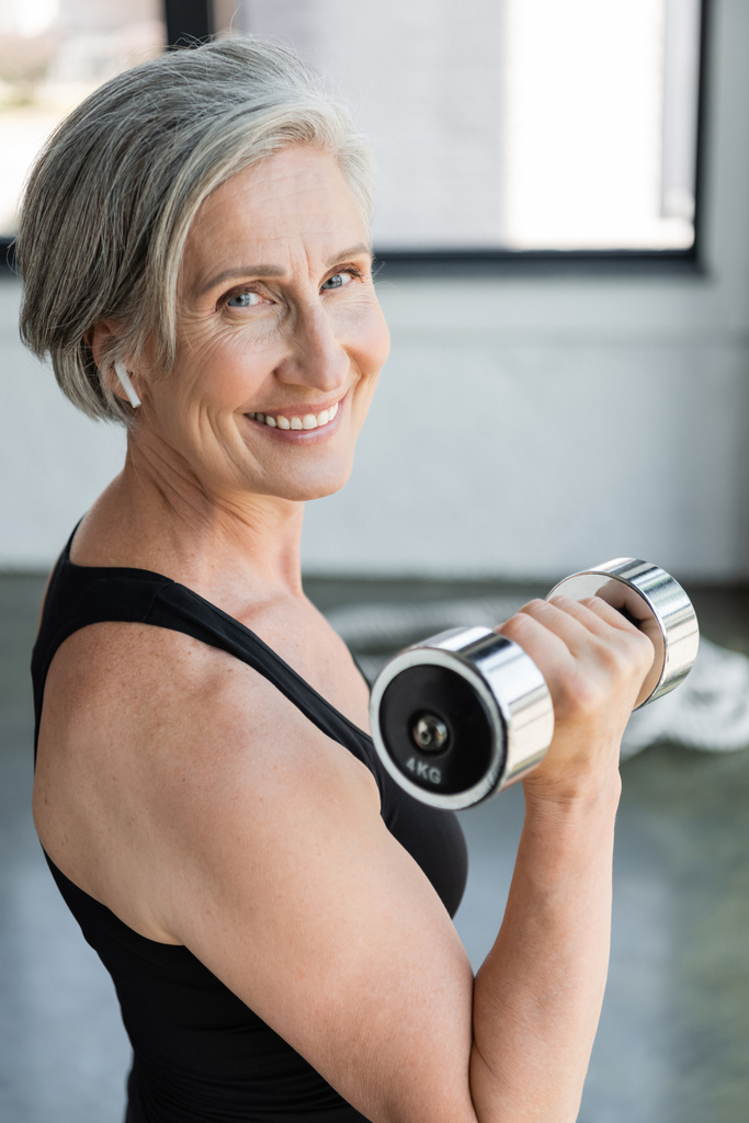 mujer mayor sonriente en auriculares inalámbricos escuchando música y haciendo ejercicio con mancuerna en el gimnasio  - Foto, Imagen
