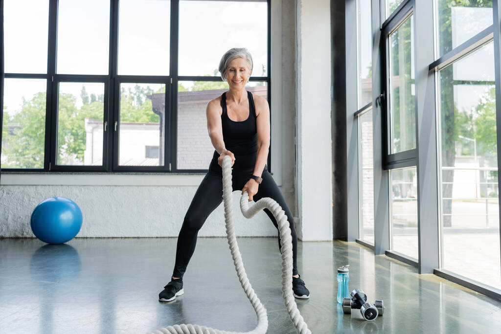 повна довжина щасливої старшої жінки, що працює з бойовими мотузками біля гантелів у спортзалі
  - Фото, зображення