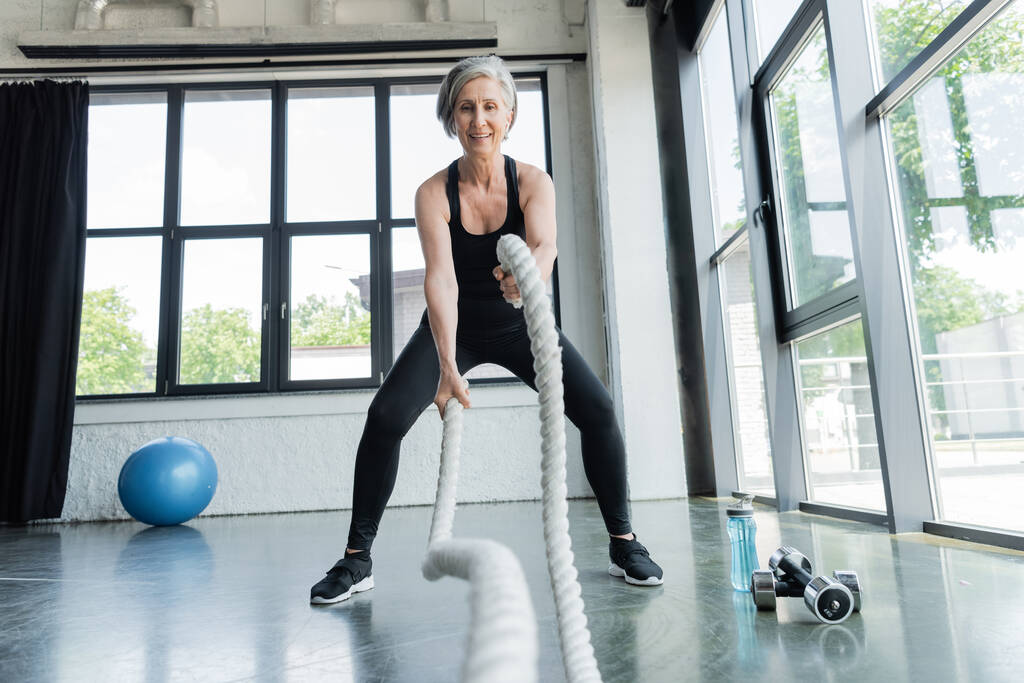 longitud completa de entrenamiento positivo de la mujer mayor con cuerdas de batalla cerca de pesas en el gimnasio  - Foto, Imagen