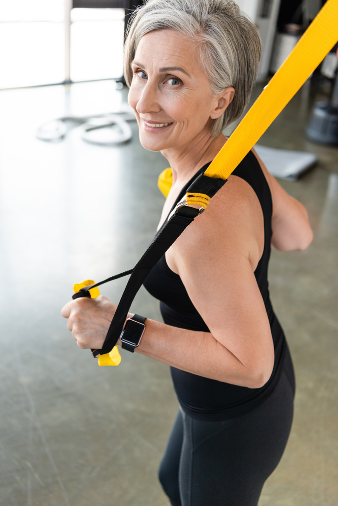 mujer mayor feliz con pelo gris haciendo ejercicio con correas de suspensión en el centro deportivo  - Foto, Imagen
