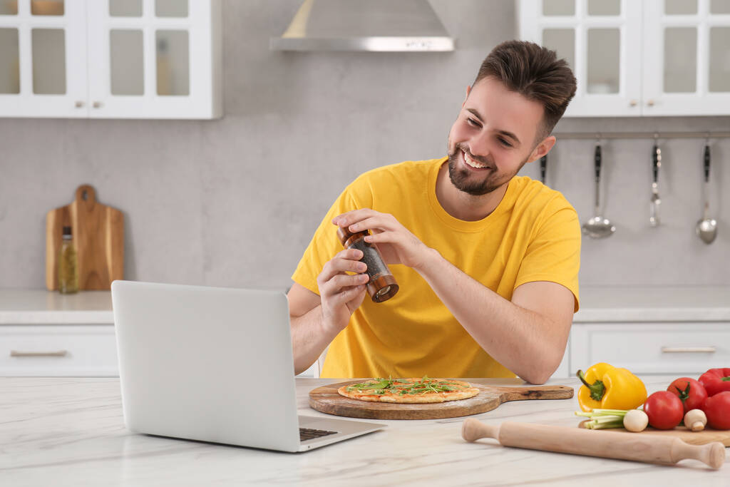 Szczęśliwy człowiek dodając pieprz na pizzy podczas oglądania gotowania online kurs w kuchni. Czas na hobby - Zdjęcie, obraz