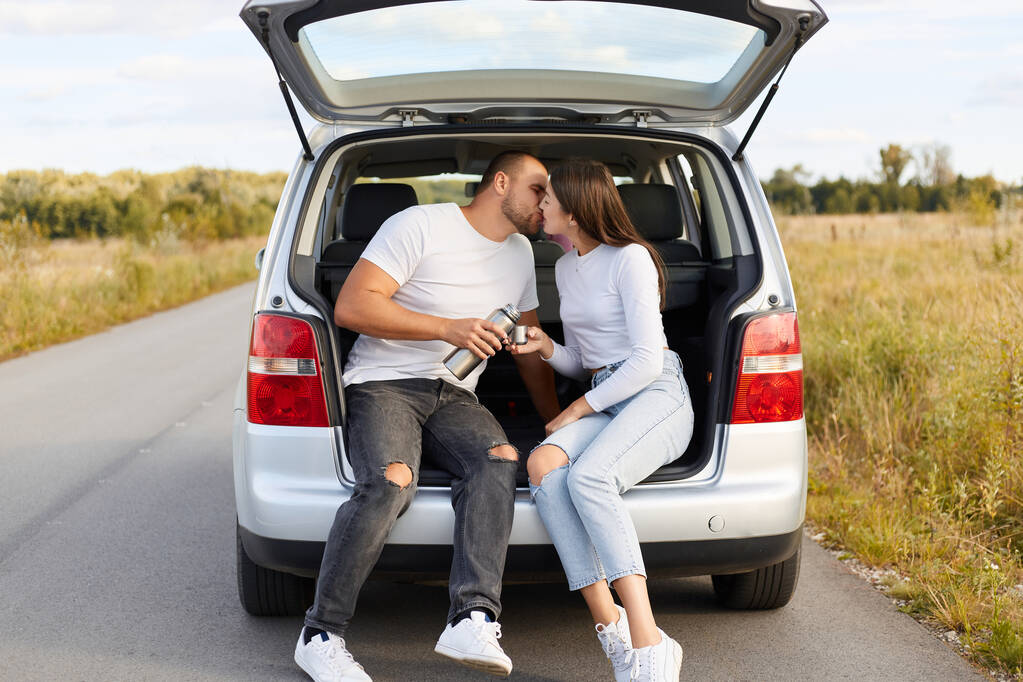 Portrait of romantic couple sitting and kissing in car trunk on road, man and woman wearing white shirts traveling together around the country, having romantic journey, honey moon by automobile. - Photo, Image