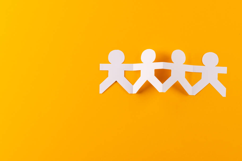 Close up of four paper cut out people figures holding hands with copy space on orange background. Humanitarian, people, help and human concept. - Photo, Image