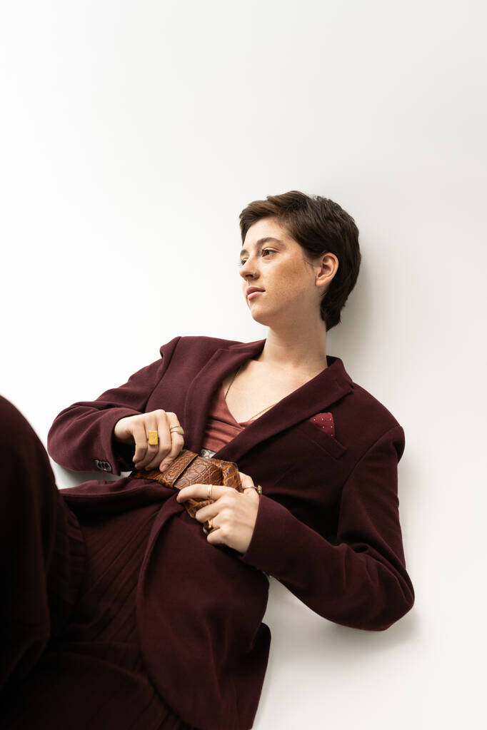 mulher sonhadora em terno elegante tocando cinto de couro e olhando para o fundo cinza - Foto, Imagem