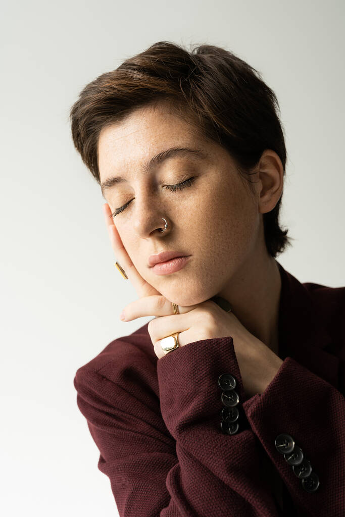 Porträt einer jungen sommersprossigen Frau in braunem Blazer, die mit geschlossenen Augen auf grau isoliert posiert - Foto, Bild