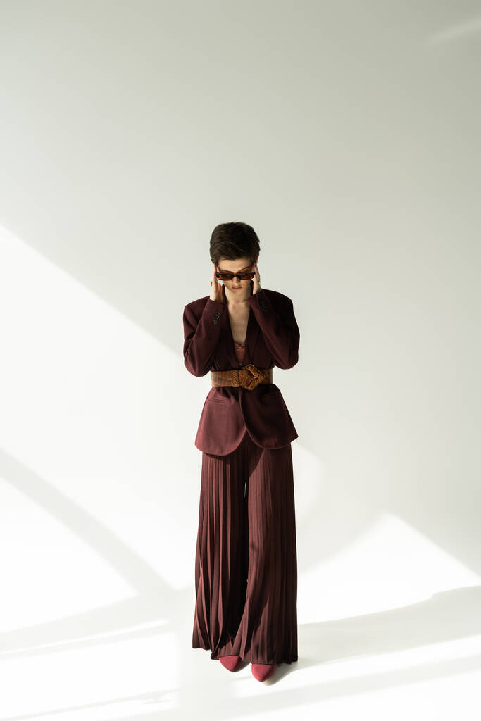 modèle brune pleine longueur en veste élégante et pantalon palazzo ajustant les lunettes de soleil sur fond gris avec éclairage - Photo, image