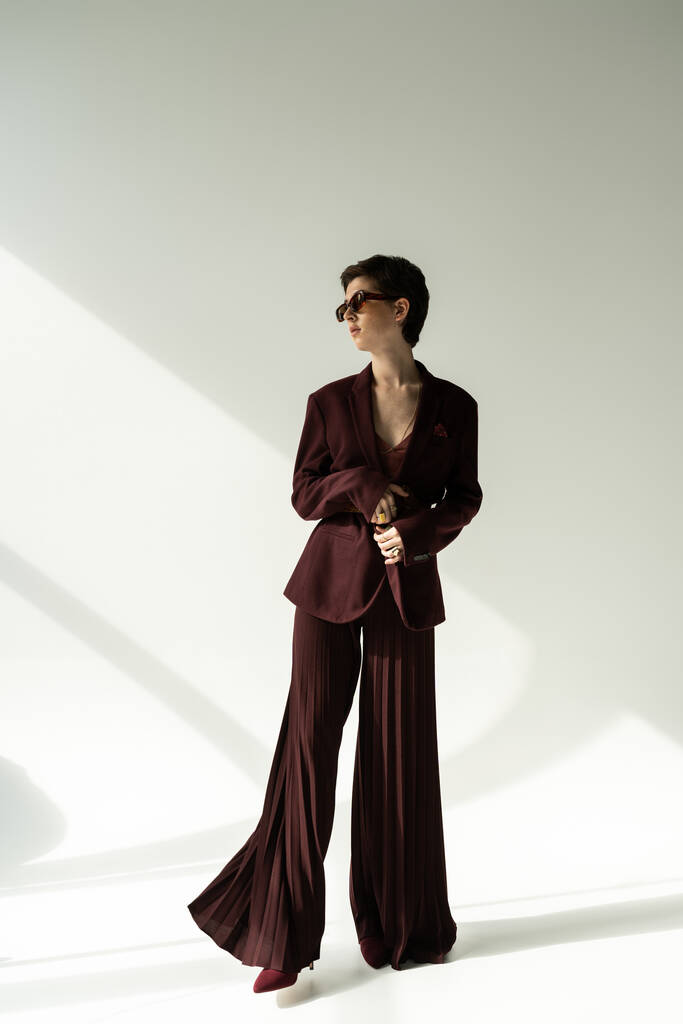 piena lunghezza di donna bruna in occhiali da sole ed elegante pantalone sfondo grigio con illuminazione - Foto, immagini