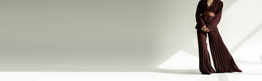 cropped άποψη του μοντέλου σε palazzo παντελόνι και σακάκι με δερμάτινη ζώνη σε γκρι φόντο με φωτισμό, πανό - Φωτογραφία, εικόνα
