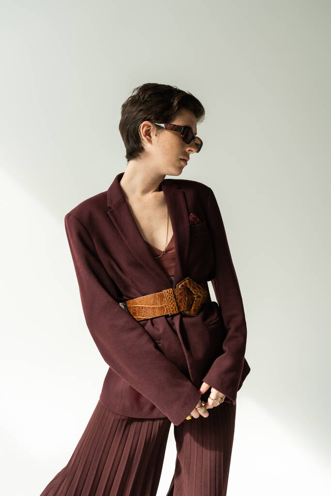 brunette woman in sunglasses and pantsuit with leather belt on grey background  - Photo, Image