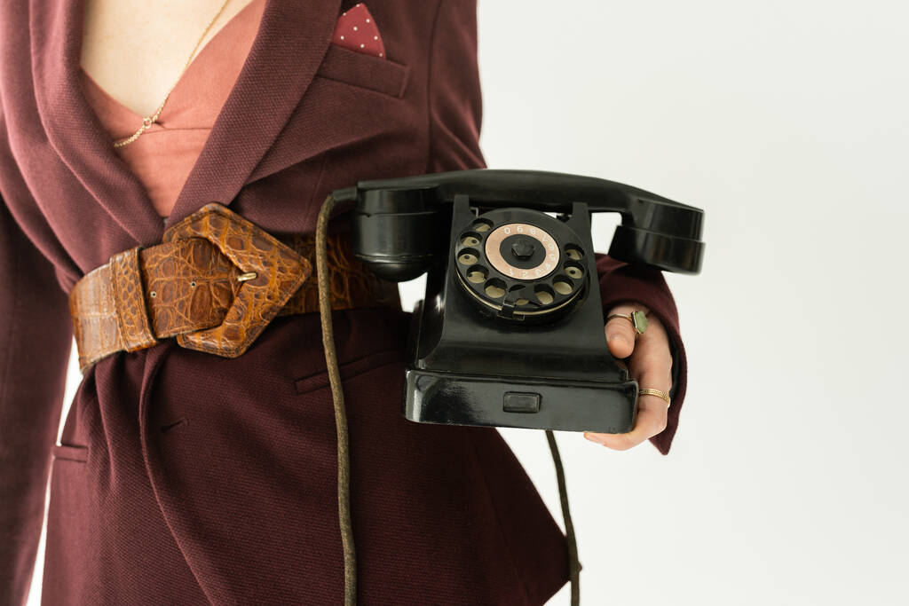 vista cortada de mulher em blazer na moda e cinto de couro segurando telefone vintage isolado em cinza - Foto, Imagem