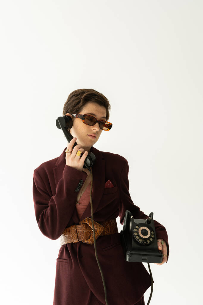 μελαχρινή μοντέλο σε γυαλιά ηλίου και μοντέρνο σακάκι με δερμάτινη ζώνη καλώντας σε vintage τηλέφωνο απομονώνονται σε γκρι - Φωτογραφία, εικόνα