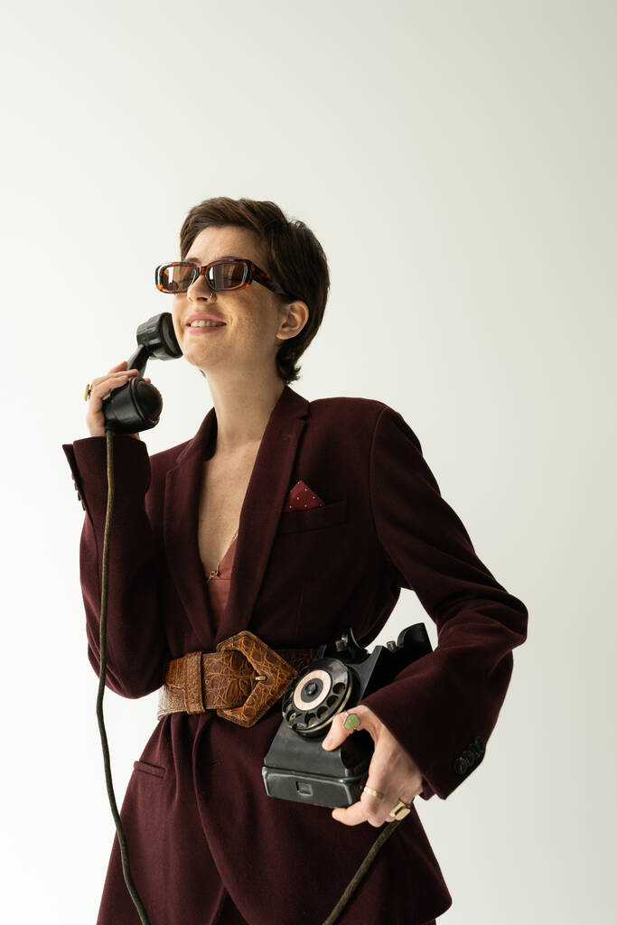 ξέγνοιαστη γυναίκα με γυαλιά ηλίου και καφέ σακάκι με δερμάτινη ζώνη που μιλάει σε ρετρό τηλέφωνο απομονωμένο σε γκρι - Φωτογραφία, εικόνα